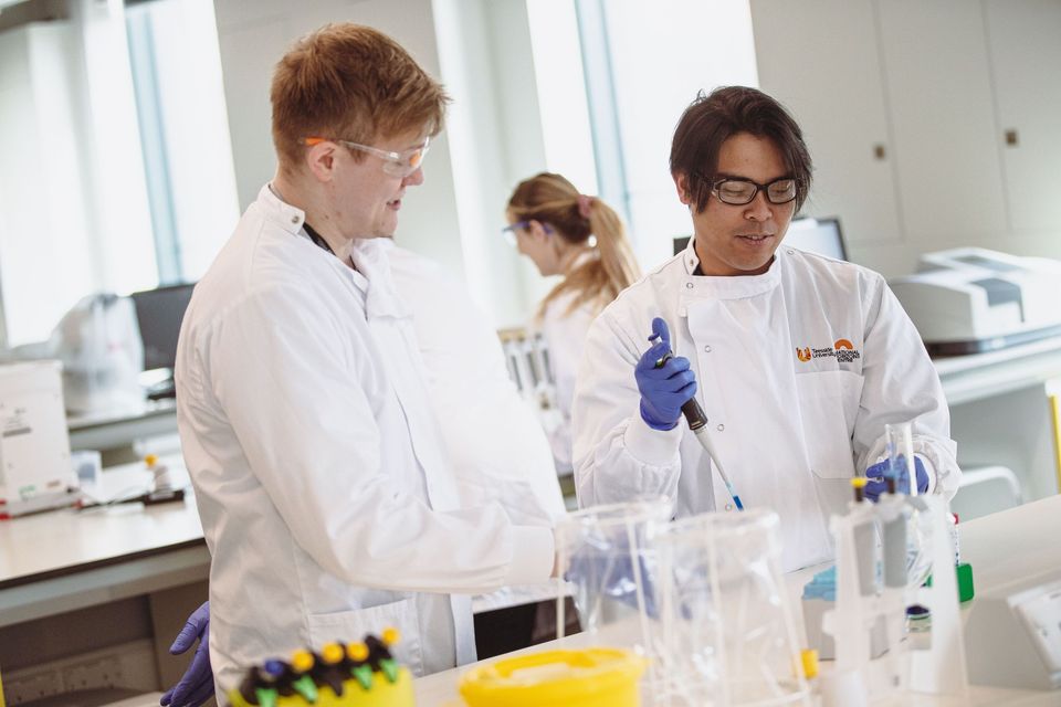 Two scientists wearing lab coats and safety goggles work together with lab equipment in a laboratory.